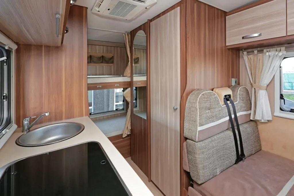 A motorhome interior looking through the kitchen bench to a bunk bed and a belted travel seat beside the bathroom door